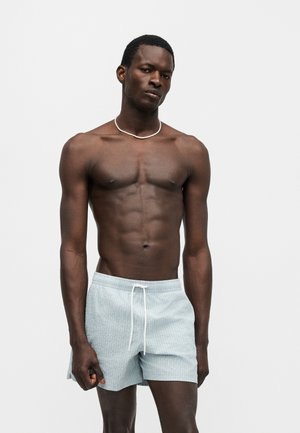 Αθλητικός μελαμψός άνδρας που φοράει ανοιχτόχρωμο μπλε μαγιό σορτς με λευκό κορδόνι και λεπτό λευκό κολιέ, στέκεται μπροστά σε ένα απλό φόντο.