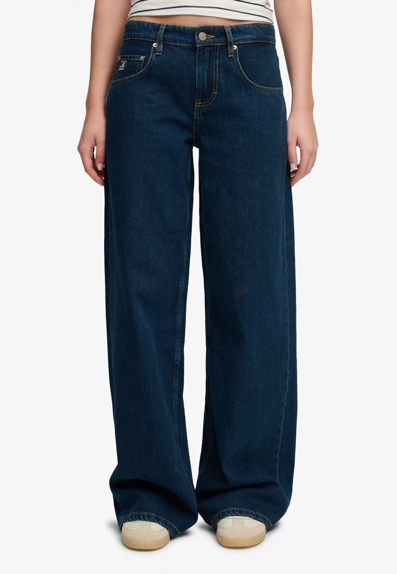 Mørkeblå straight-leg denimjeans iført beige sko, vist fra taljen til fødderne mod en hvid baggrund.