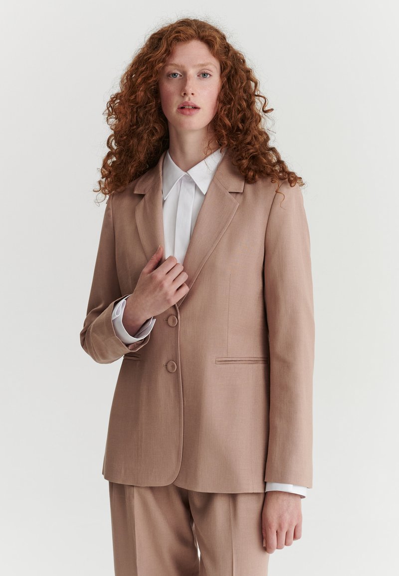 Femme aux cheveux roux bouclés, portant une veste et un pantalon beige sur une chemise blanche à col, debout devant un fond clair uni.