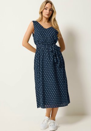 Frau trägt ein ärmelloses, marineblaues Midikleid mit kleinen weißen rechteckigen Mustern, einem gebundenen Taillengürtel und weißen Sneakern, steht vor einer schlichten Wand.