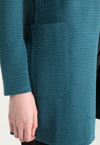 Tyrkysová texturovaná bunda s lehce volným střihem, která má na přední straně náprsní kapsu a rukávy o délce tří čtvrtin.