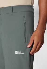 Vihreät kevyet housut, joissa on vetoketjullinen tasku ja valkoinen logo "Jack Wolfskin" reidessä. Sujuva pinta ja istuva muotoilu.