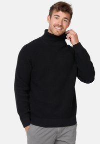 Maglione a collo alto nero con un pattern a maglia testurizzato, caratterizzato da polsini e orlo a costine, indossato da un modello sorridente.