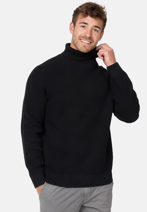Suéter negro de cuello alto con un patrón de tejido texturizado, con puños y dobladillo de acanalado, exhibido en un modelo sonriendo.