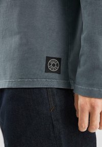 Graues Langarmshirt mit weicher Textur, ausgestattet mit einem schwarzen quadratischen Etikett mit "QUITE SENSE" nahe dem Saum. Trägt es über einer dunklen Jeans.