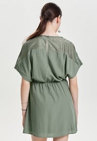 Vestido corto verde con corte suelto, que presenta un patrón de encaje texturizado en los hombros, cintura elástica y un detalle de abertura en la espalda.