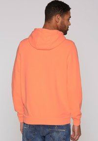 Sudadera con capucha de tela naranja brillante, con un bolsillo tipo canguro, puños y dobladillo acanalados. El diseño es simple y relajado.