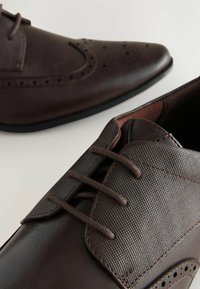 Chaussures en cuir marron avec un dessus texturé, présentant un petit perforé détaillé, cinq œillets et une semelle contrastante dans un design élégant.