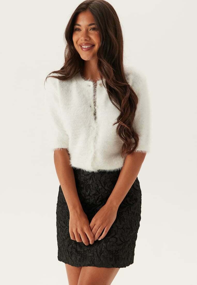 Donna sorridente, che indossa un cardigan corto e morbido bianco con bottoni e una gonna nera a pieghe fino al ginocchio.