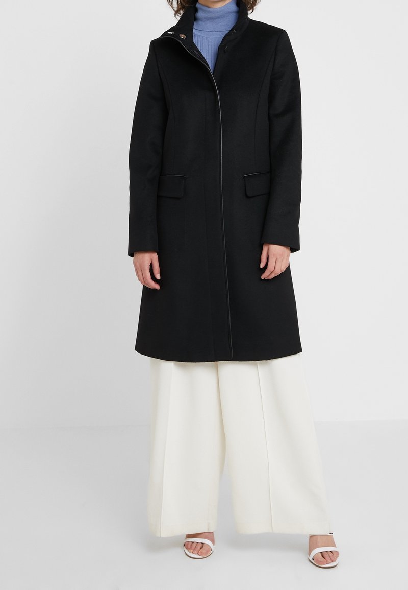 Personne portant un long manteau noir par-dessus un col roulé bleu, un pantalon blanc large et des sandales blanches à bout ouvert sur un fond uni.