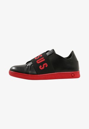 Zapatilla negra de corte bajo con una amplia correa negra con letras rojas "SUS" y suela y ribete de color rojo brillante.