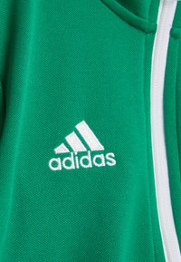 Survêtement en tissu vert avec une surface texturée. Fermeture éclair blanche et logo Adidas brodé en blanc sur le côté gauche.