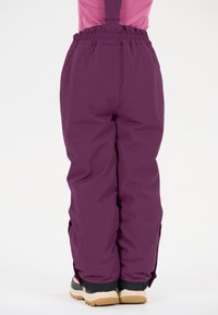 Lila Schneehosen mit verstellbaren Trägern, elastischem Bund und Reißverschlüssen an den Knöcheln, kombiniert mit schwarzen und beigen Winterstiefeln.