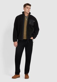 Zwarte fleece jas met hoge kraag, ritssluiting aan de voorkant en een patch zak. Draagt over een groene trui, in combinatie met zwarte broek en bruine schoenen.