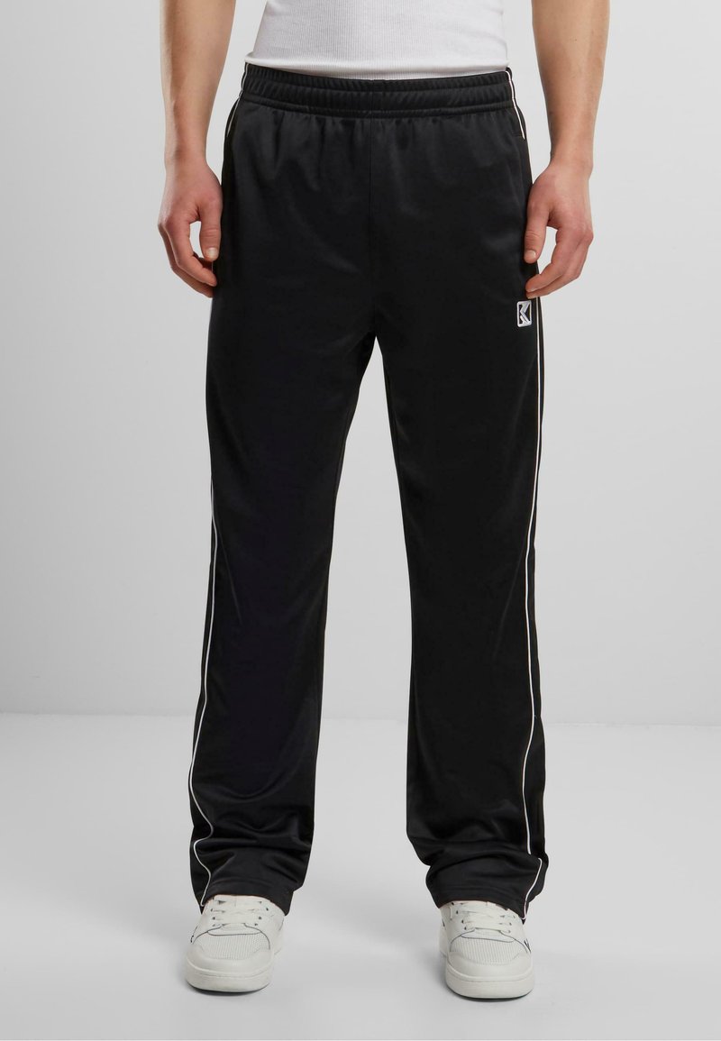 Pantaloni atletici negri cu un finisaj mat, având dungi albe pe laterale, talie elastică și un mic logo pe coapsa stângă.