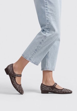 Piernas con vaqueros cortos azul claro y zapatos Mary Jane planos color marrón oscuro con tachuelas y hebilla caminando sobre un fondo claro y liso.