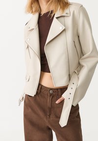 Giacca biker corta beige con hardware argentato, cerniera asimmetrica e dettagli con bottoni a pressione. Abbinata a pantaloni in velluto a coste marroni.