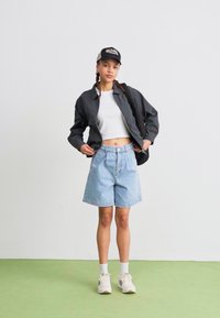 Fekete cipzáros dzseki, fehér cropped felső, világoskék farmer rövidnadrág, fehér zoknik és sportcipők. A modellen fekete sapka van. Egyszínű háttér.