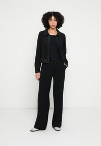 Jachetă neagră cu șnur, combinată cu un top negru și pantaloni cu picior lejer. Textură netedă; design minimalist; completat de pantofi negri și albi.