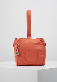 Mała koralowa torebka z górnym paskiem, okrągłą łatką z logo oraz małym srebrnym breloczkiem w kształcie małpy na gładkiej białej powierzchni.