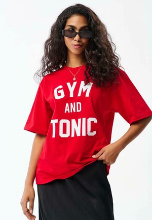 Kobieta w czarnych okularach przeciwsłonecznych i czerwonej koszulce z dużym białym napisem „"GYM AND TONIC,"” pozuje z jedną ręką na biodrze.