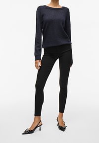 Pull en maille bleu marine à col rond, associé à un pantalon noir ajusté. Des talons noirs à bout pointu avec un nœud complètent la tenue.