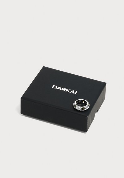 Boîte rectangulaire noire avec texte blanc "DARKAI" et une bague en argent avec des pierres sombres reposant sur son coin.