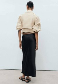 Veste beige cropped avec des manches bouffantes, associée à une élégante jupe maxi noire. Le mannequin porte des sandales noires simples, sur un fond neutre.