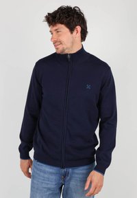 Sweat à zip bleu marine avec un col montant, fabriqué en tissu tricoté. Il présente un petit logo sur la poitrine gauche et des poignets et un ourlet côtelés.