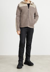 Giacca marrone e beige con zip e cappuccio, texture in lana e design a pannelli contrastanti; abbinata a pantaloni neri e scarpe nere.