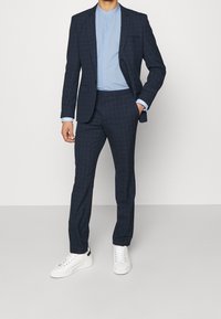 Man in een marineblauw geruit pak met bijpassende broek, lichtblauwe shirt en witte sneakers, staande tegen een effen achtergrond.