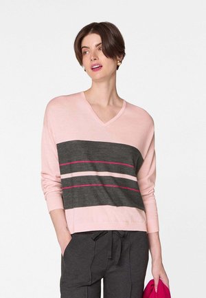 Vrouw met een roze en donkergrijs gestreepte V-hals trui en donkergrijze broek, die in haar linkerhand een opgevouwen roze kledingstuk vasthoudt.