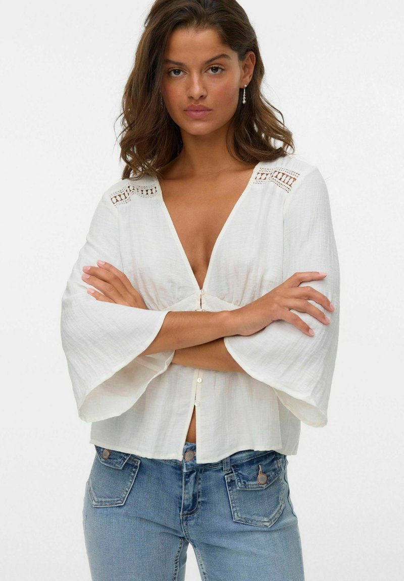 Blusa bianca leggera con scollatura a V profonda, ampie maniche a campana e dettagli in pizzo decorativi alle spalle. Chiusura con bottoni davanti.