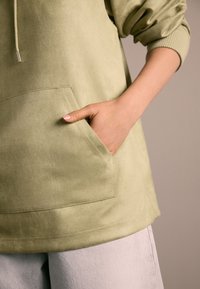 Olivovo zelená mikina s jemnou textúrou, predným vreckom, sťahovacou šnúrkou a rebrovanými manžetami. Kombinovaná so svetlošedými nohavicami.