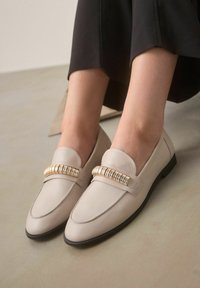 Beige nahka loaferit pyöreällä varpaalla, joissa on koristeellinen kultasävyinen metalliyksityiskohta varpaankärjessä ja litteä musta pohja.
