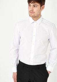 Weißes Langarmhemd mit spitzen Kragen, Knopfleiste vorne und geknöpften Manschetten, kombiniert mit schwarzen Hosen aus glattem Stoff.