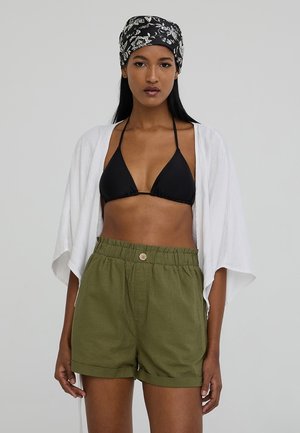 Top bikini nero, copertura bianca aperta davanti e pantaloni corti verdi oliva con cintura elasticizzata e dettaglio bottone. Tessuti lisci, vestibilità comoda.