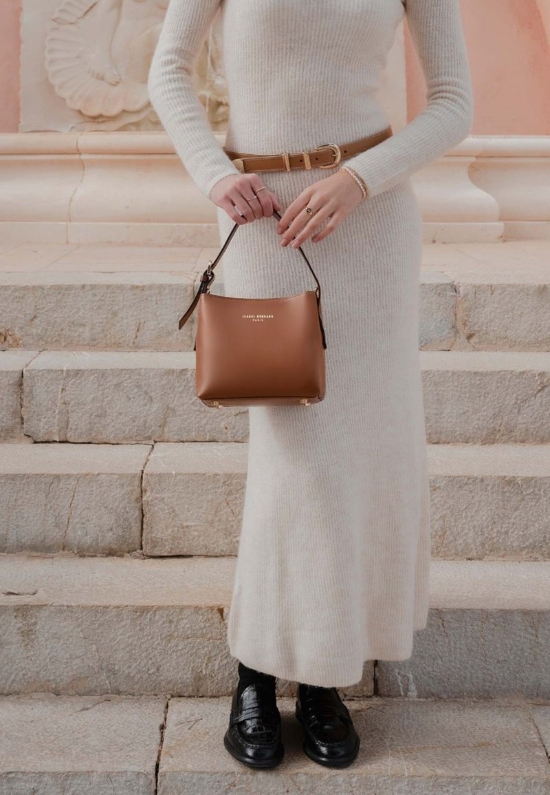 Personne en robe tricotée crème avec une ceinture beige, tenant un sac à main marron, debout sur des marches en pierre. Chaussures noires et détails d’arrière-plan visibles.