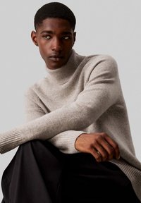 Maglione a collo alto grigio chiaro con polsini e orlo a coste, indossato sopra un tessuto scuro. Texture liscia con un sottile motivo a maglia.