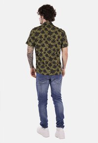 Maglietta a maniche corte con base verde e motivo di palme nere, abbinata a jeans blu e sneakers bianche. Tono della pelle visibile sulle braccia.