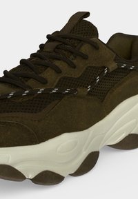 Gros plan sur une basket vert foncé avec des lacets noirs, une texture en mesh et en suède, un lacet blanc à motifs et une semelle blanche épaisse.