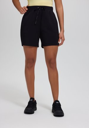 Kobieta ubrana w czarne szorty z troczkiem i czarne buty sportowe, stojąca na tle jednolitego jasnego tła.