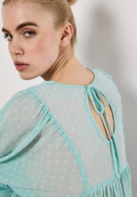 Eine hellblaue, transparente Bluse mit strukturiertem Muster, V-Ausschnitt hinten und einer Schleifenverschluss. Mit Rüschensaum entlang der Ärmel.