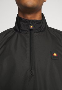 Pull-over noir à fermeture quart-zip avec col montant, comportant un écusson logo coloré et un tissu texturé. Design minimaliste avec une finition lisse.