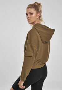 Sudadera corta de color verde oliva con capucha ajustable, bolsillo lateral y puños acanalados. Combinada con leggings negros. Textura de tela suave.
