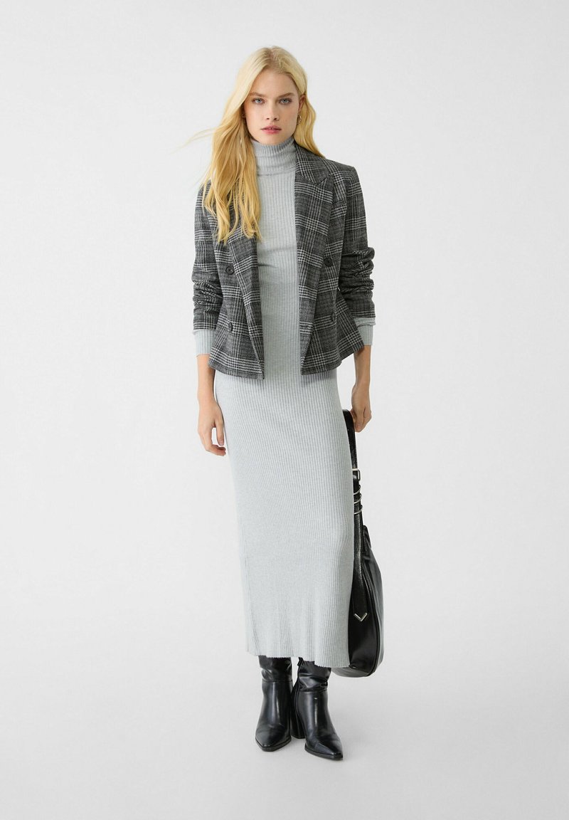 Mujer rubia vistiendo un vestido gris de cuello alto acanalado, un blazer de cuadros, botas negras y sosteniendo un bolso negro frente a un fondo blanco liso.