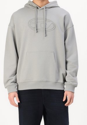 Sweat à capuche gris clair avec poche avant, logo embossé subtil, porté par une personne en pantalon foncé, visible du cou aux hanches.