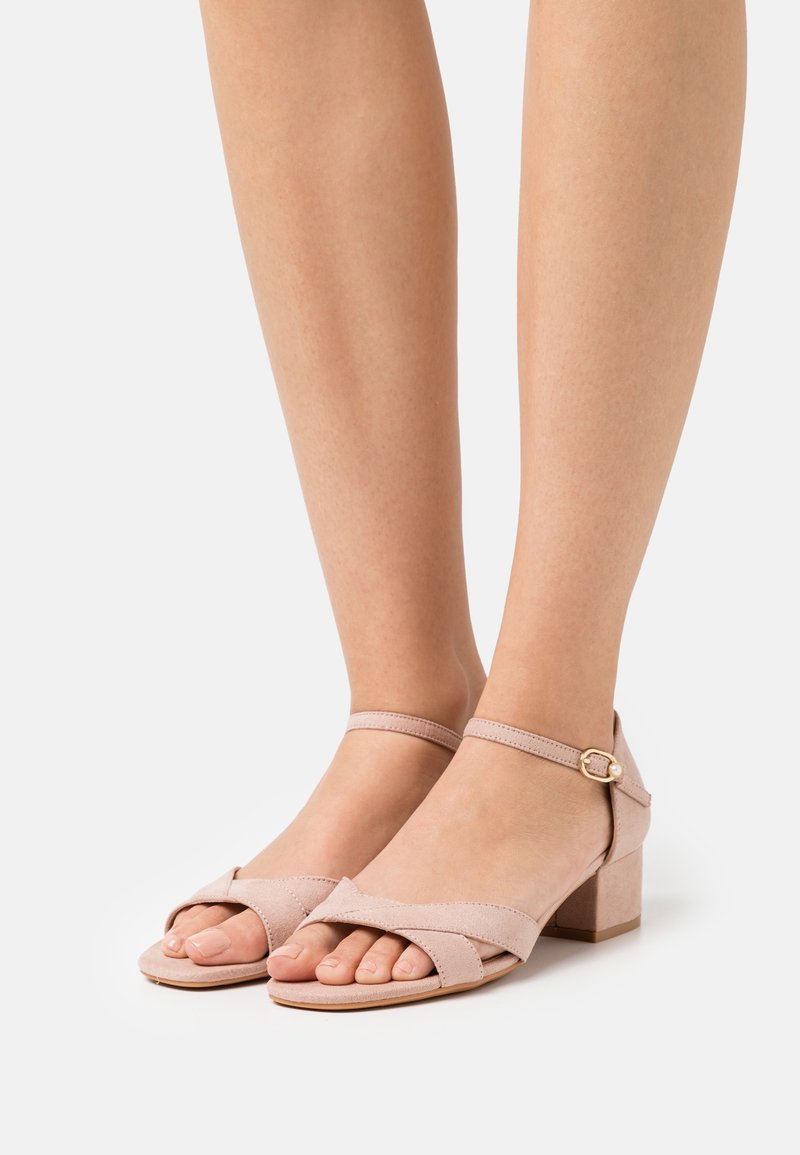 Pieds portant des sandales nude roses en daim avec talons carrés, lanières à la cheville et orteils ouverts sur fond blanc.
