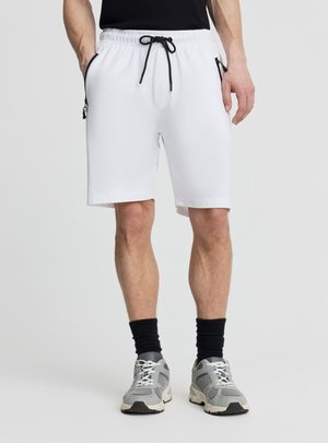 Uomo che indossa pantaloncini bianchi con coulisse e tasche con cerniera, calzini neri, scarpe da ginnastica grigie e maglietta nera, in piedi con le mani nelle tasche.