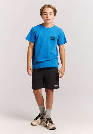 Blauw katoenen T-shirt met zwarte grafische print, gecombineerd met zwarte shorts en grijze sokken. De outfit bevat casual sneakers met een patroon.